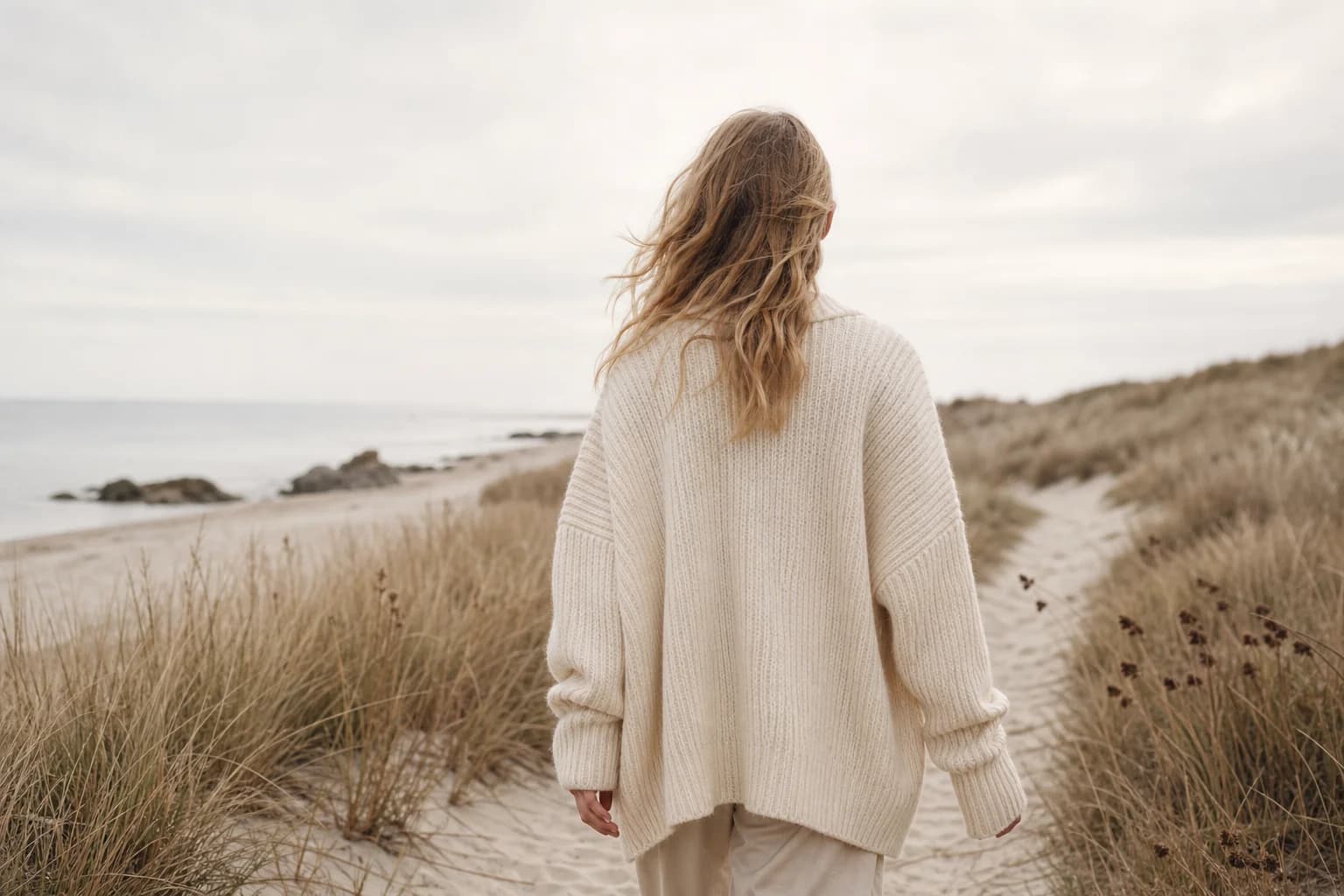 A woman in an ivory ribbed cardigan walking along a quiet coastal path